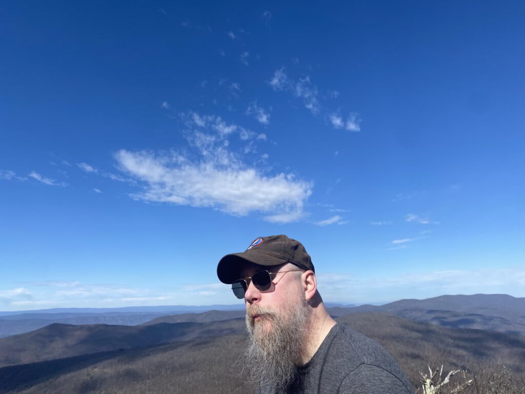 Your host, J.M. Hart with a substantial vista of Blue Ridge mountains behind him. The sky is blue with a few light, white, clouds. 